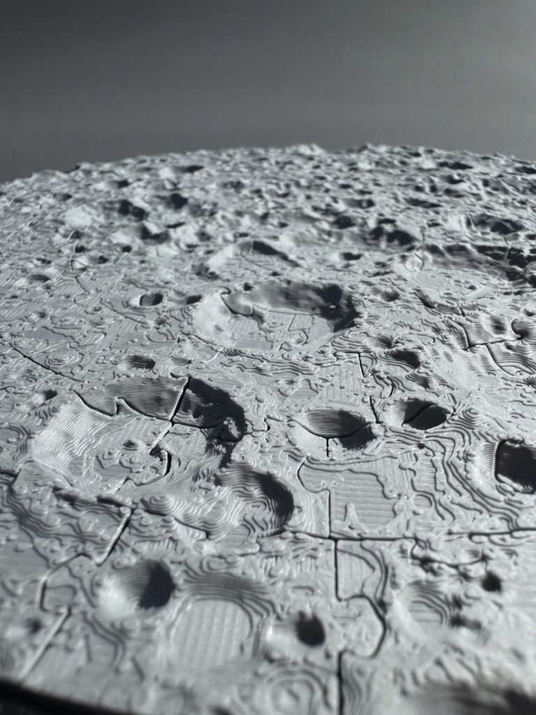 A close-up in low, raking light of printed crater relief in gray filament, with the puzzle cuts running between the craters